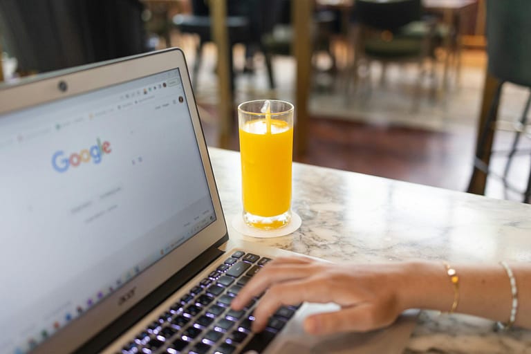 Woman typing on laptop with Google homepage open, beside a glass of orange juice on a marble table.